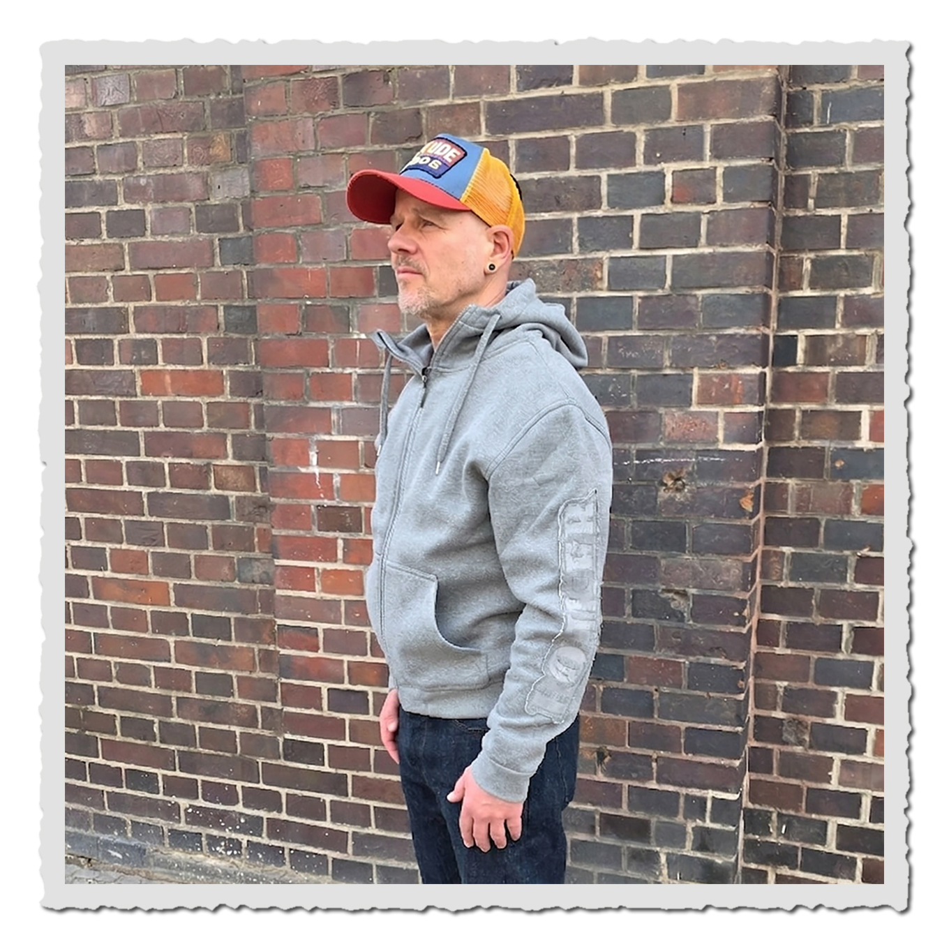 Person in hellgrauem Zip Hoodie in Seitenansicht vor Backsteinwand, mit bunter Kappe und dunkler Jeans.